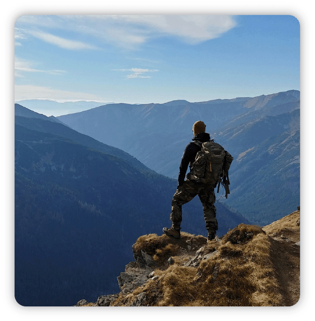 Hiker on mountain peak with back turned, looking into the horizon at other mountain ranges set against blue skies and clouds