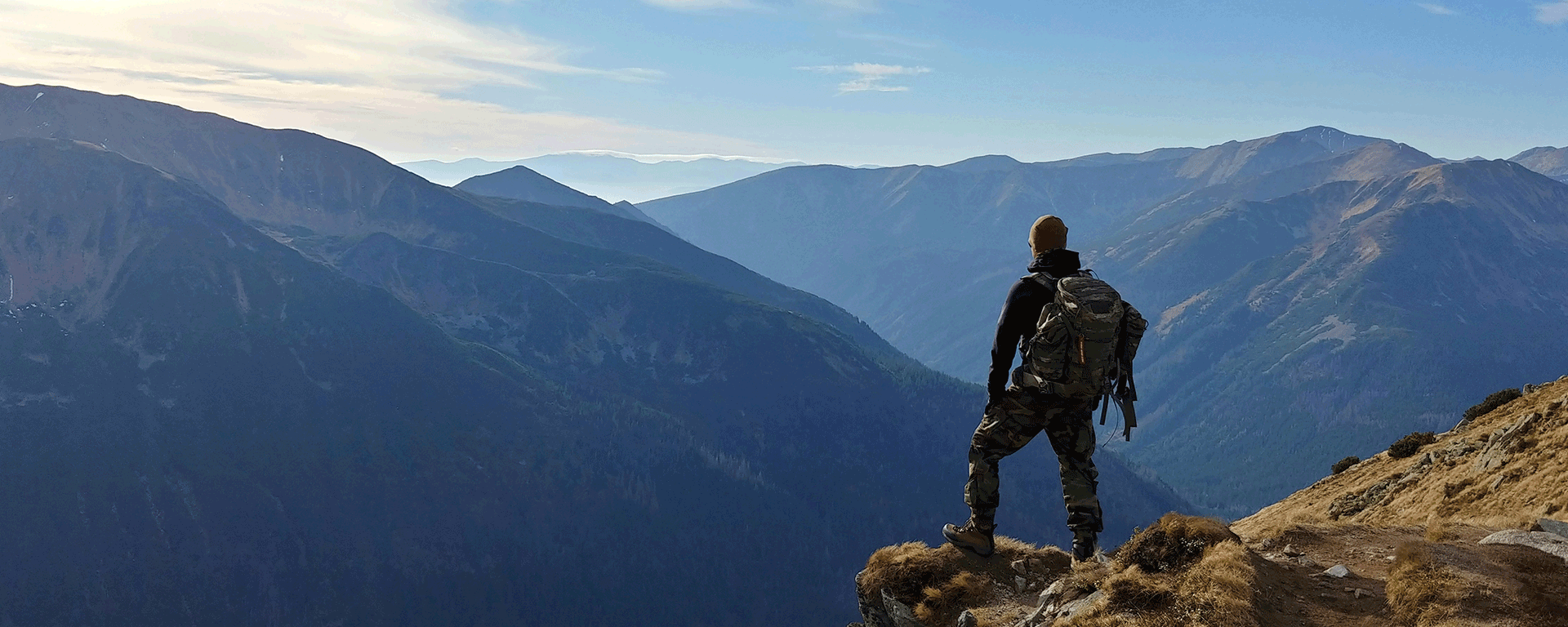 Hiker on top of a mountain looking out at mountain ranges set against blue sky and clouds