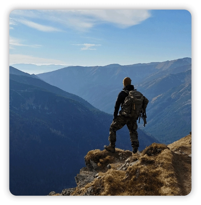 Hiker on mountain peak with back turned, looking into the horizon at other mountain ranges set against blue skies and clouds
